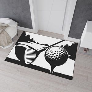 以下が含まれることがあります： ゴルフをテーマにした白黒のドアマット。ゴルフのクラブとティーに乗ったゴルフボールが描かれています。デザインには、背景に木々のあるゴルフコースの様式化された描写が含まれています。マットは長方形です。