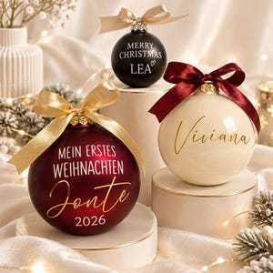 May include: Three round Christmas ornaments with ribbons. One matte black with 'MERRY CHRISTMAS LEA' and a heart, one cream with 'Viviana' in gold script and a dark red ribbon, and one deep red with 'MEIN ERSTES WEIHNACHTEN Jonte 2026' in gold.