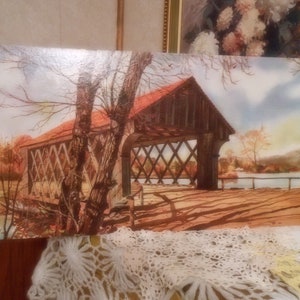 May include: A framed landscape painting depicts a covered bridge with a red roof, set against a backdrop of autumn foliage and a calm river. The bridge's wooden structure is detailed, with shadows cast across the wooden planks.