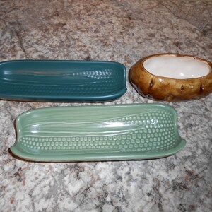 May include: Three ceramic serving dishes: two corn-shaped in teal and light green, and a potato-shaped dish in brown with a white interior. The corn dishes have detailed textures.