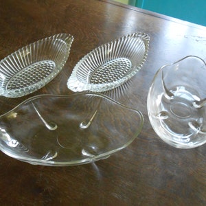 Pode incluir: Quatro tigelas de vidro transparente de formas e tamanhos diferentes. Duas das tigelas são de forma oval com um design nervurado. Uma tigela é redonda com uma borda ondulada. A quarta tigela é de forma oval com uma superfície lisa.