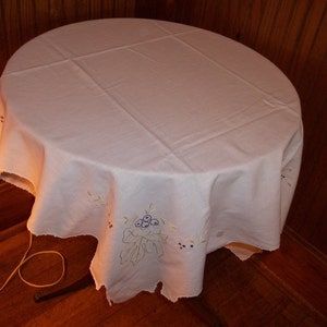 May include: White embroidered tablecloth with a floral design featuring a cluster of purple berries and green leaves. The tablecloth is draped over a round table.