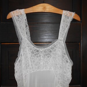 K&ouml;nnte beinhalten: Ein wei&szlig;es Nachthemd im Vintage-Stil mit zarten Spitzendetails an Oberteil und Tr&auml;gern. Das Kleid h&auml;ngt an einem Holzkleiderb&uuml;gel vor einer dunklen Holzt&uuml;r. Das Nachthemd ist ein klassisches W&auml;schest&uuml;ck.