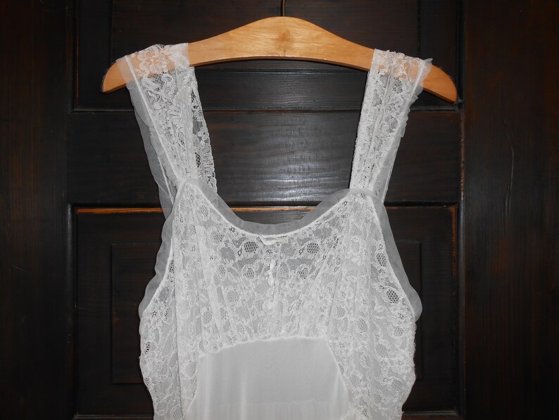 K&ouml;nnte beinhalten: Ein wei&szlig;es Nachthemd im Vintage-Stil mit zarten Spitzendetails an Oberteil und Tr&auml;gern. Das Kleid h&auml;ngt an einem Holzkleiderb&uuml;gel vor einer dunklen Holzt&uuml;r. Das Nachthemd ist ein klassisches W&auml;schest&uuml;ck.