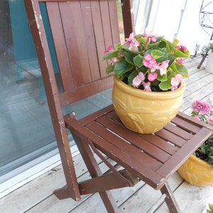 May include: A brown wooden folding chair with a yellow planter filled with pink flowers on the seat. The chair is on a wooden deck.