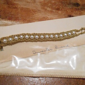 May include: A gold chain with white pearls, labeled "Sweater Guard" on a white card.