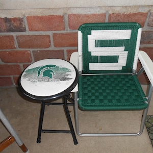 May include: A green and white folding chair with a white stripe pattern on the back. A black folding stool with a white circular seat featuring the Michigan State Spartan logo and the words "Michigan State".