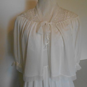 K&ouml;nnte beinhalten: Elfenbeinfarbenes Vintage-Dessous-Set mit einem langen Nachthemd und einer passenden transparenten Jacke. Die Jacke hat zarte Spitzendetails an den Schultern und einen Bindverschluss. Die &Auml;rmel sind dreiviertellang mit ger&uuml;schten Manschetten.