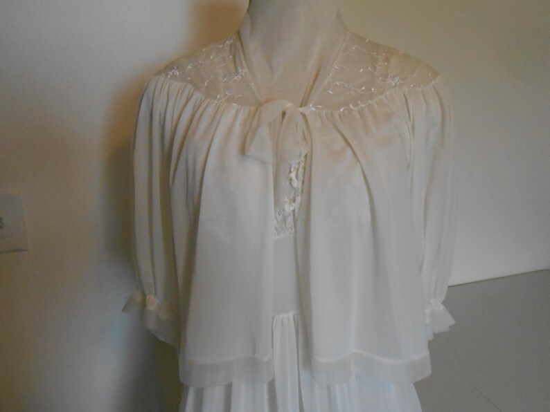 K&ouml;nnte beinhalten: Elfenbeinfarbenes Vintage-Dessous-Set mit einem langen Nachthemd und einer passenden transparenten Jacke. Die Jacke hat zarte Spitzendetails an den Schultern und einen Bindverschluss. Die &Auml;rmel sind dreiviertellang mit ger&uuml;schten Manschetten.
