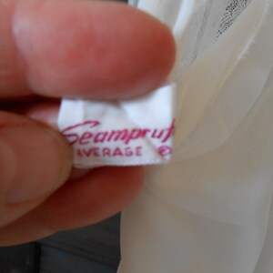 K&ouml;nnte beinhalten: Nahaufnahme eines wei&szlig;en Etiketts mit dem Wort "Seamproof" in rosa Schrift. Das Wort "AVERAGE" ist darunter in Rosa gedruckt, mit einem kleinen Symbol rechts. Das Etikett wird in einer Hand gehalten, mit einem durchsichtigen, cremefarbenen Stoff im Hintergrund.