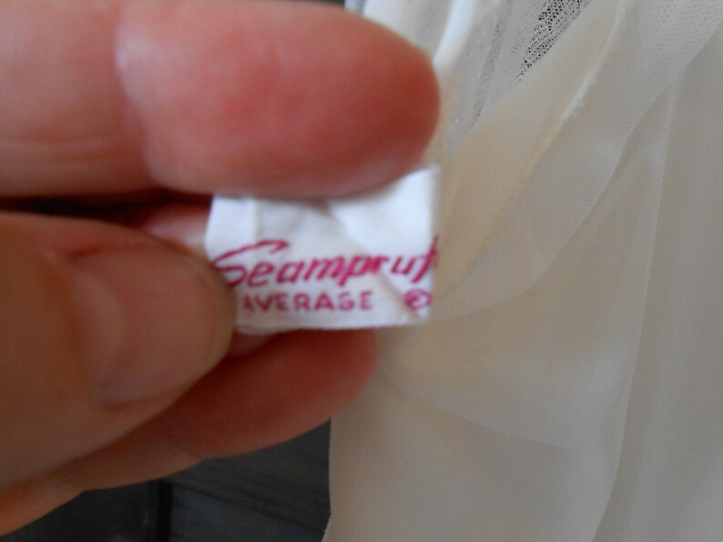 K&ouml;nnte beinhalten: Nahaufnahme eines wei&szlig;en Etiketts mit dem Wort "Seamproof" in rosa Schrift. Das Wort "AVERAGE" ist darunter in Rosa gedruckt, mit einem kleinen Symbol rechts. Das Etikett wird in einer Hand gehalten, mit einem durchsichtigen, cremefarbenen Stoff im Hintergrund.