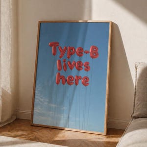 Könnte beinhalten: Ein gerahmter Kunstdruck mit einem blauen Himmel als Hintergrund und roten Ballonbuchstaben, die "Type-B lives here" schreiben. Das Kunstwerk befindet sich in einem Holzrahmen und lehnt an einer Wand. Das Bild ist gut beleuchtet und hat eine moderne Ästhetik.