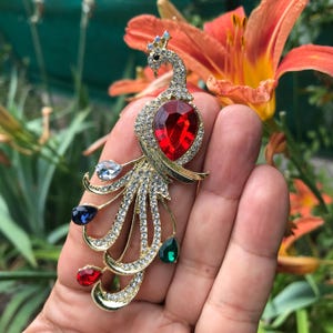Peut inclure: Une broche paon dorée ornée d'une grande pierre centrale rouge en forme de goutte, de cristaux pavés transparents sur le corps et la queue, et de quatre pierres d'accent en forme de goutte transparentes, bleu foncé, vertes et rouges.