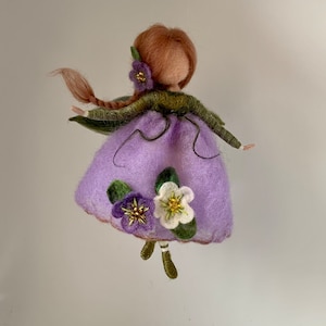 Könnte beinhalten: Eine nadelgefilzte Puppe mit rotbraunem Haar in einem Zopf, die ein lavendelfarbenes Kleid mit grünen Ärmeln und einem dunkelgrünen Oberteil trägt. Das Kleid ist mit einer violetten und einer weißen Blume verziert. Die Puppe hat grüne Beine und kleine Flügel.