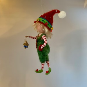 Può includere: Un ornamento per l'albero di Natale in feltro a forma di elfo con un cappello rosso, pantaloni a righe verdi e bianche e un ornamento a campana dorata in mano.