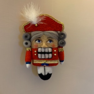 Può includere: Un ornamento schiaccianoci in feltro con uniforme rossa e finiture dorate, una piuma bianca su un cappello rosso, capelli grigi ricci e occhi blu. La figura ha un corpo rettangolare con base bianca e nera, bottoni dorati e polsini bianchi.
