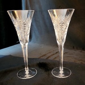Peut inclure: Deux flûtes à champagne en cristal transparent avec des motifs floraux gravés. Les flûtes ont de longues tiges fines et des coupes coniques larges. Les bases sont rondes. Les verres sont présentés sur fond sombre.