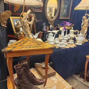 May include: A display of antique items, including a small wooden table with a decorative top, brown leather boots, and a collection of teacups and teapots. The scene is illuminated by lamps with fringed shades, creating a vintage aesthetic.