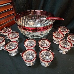 May include: A vintage punch bowl set with a large, clear glass bowl, a ladle, and twelve matching cups. The set features a red trim and a diamond pattern. The cups have handles and sit on clear glass bases.