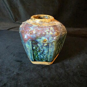 May include: A vintage, hexagonal-shaped ceramic vase with a wide opening. The vase features a blend of colors, including blue, brown, and hints of green. The exterior has a textured appearance, suggesting an aged or weathered finish. The vase is likely a decorative item.