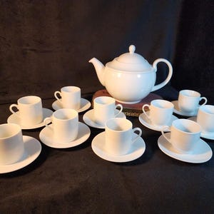 May include: A white porcelain tea set, including a teapot and several teacups with saucers. The set is arranged on a dark surface, creating a classic and elegant aesthetic. The cups have handles and are designed for tea service.