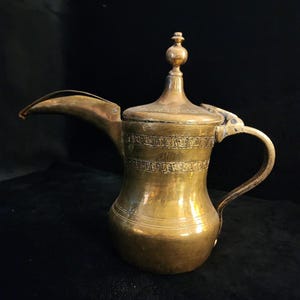 May include: A vintage brass coffee pot with a long spout, a handle, and a decorative lid. The pot has etched horizontal bands around the body and a small finial on top. The overall design suggests an antique or Middle Eastern style.