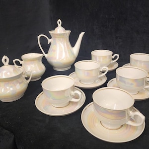 Puede incluir: Un juego de té de porcelana iridiscente vintage. El juego incluye una tetera, azucarero, lechera y ocho tazas de té con platillos. Las piezas tienen un acabado nacarado con ribetes dorados, creando una estética clásica y elegante.