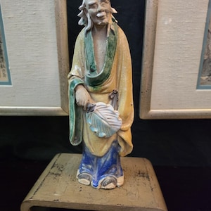 Op de afbeelding: Een keramieken beeldje van een oude man in traditionele kleding. De figuur draagt een gele jurk met groene randen en een blauwe broek. Hij houdt een waaier vast en staat op een kleine, verouderde houten standaard. Het beeldje heeft een verweerde uitstraling.