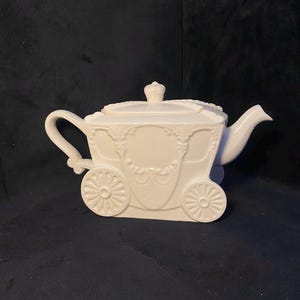 May include: A white ceramic teapot shaped like a carriage. The teapot features decorative wheels, a crown on the lid, and ornate detailing. The handle and spout are curved, and the overall design is elegant and whimsical.