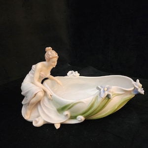 Puede incluir: Un cuenco decorativo de porcelana con una figura femenina con un vestido fluido, inclinada sobre el cuenco. El cuenco es blanco con detalles florales verdes y azules. Una pieza delicada y elegante.