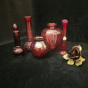 Op de afbeelding: Een verzameling robijnrode glazen vazen en decoratieve items. De collectie omvat een kleine parfumfles, een hoge getextureerde vaas, een ronde vaas, een bolvormige vaas, een slanke vaas en een decoratieve roos met groene bladeren. De items zijn geplaatst op een donkere ondergrond.