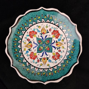 Puede incluir: Un plato decorativo de cerámica con borde festoneado. El plato presenta un borde verde azulado y un diseño central con motivos florales rojos, amarillos y azules sobre fondo blanco. El plato tiene un acabado brillante y un borde blanco.