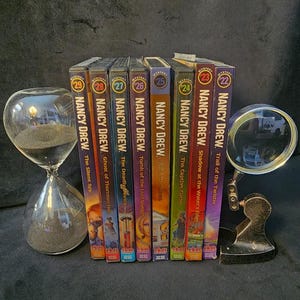 May include: A collection of Nancy Drew mystery books, including titles like "The Silent Spy" and "Trail of the Twister." The books are arranged in a row with an hourglass and a magnifying glass. The book spines are colorful.