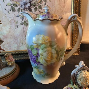 Könnte beinhalten: Vintage Porzellankrug mit Deckel, verziert mit handgemalten gelben Rosen und lila Blumen. Der Krug hat einen geschwungenen Griff und Ausguss, mit goldenen Zierleisten. Der Hintergrund ist ein Farbverlauf aus Hellblau und Pfirsich. Ein dekoratives Stück.