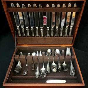 Puede incluir: Una caja de cubiertos de madera abierta, que revela un juego completo de cubiertos. El juego incluye cuchillos, tenedores y cucharas, todos con mangos ornamentados. El interior está forrado con fieltro marrón, proporcionando una presentación lujosa.