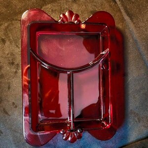 Puede incluir: Una bandeja de servicio rectangular de cristal rojo rubí con un interior dividido en una sección grande semicircular y dos compartimentos rectangulares más pequeños. La bandeja tiene un acabado liso y bordes festoneados decorativos en los extremos.