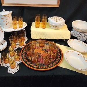 Peut inclure: Un ensemble de vaisselle en céramique blanche orné de motifs de feuilles d'automne, comprenant une carafe, des bols et des assiettes. Des verres ambrés sont disposés sur un plateau décoratif rond et à côté, avec une cocotte à couvercle.