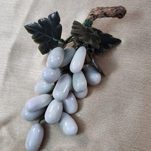 May include: A decorative sculpture of a bunch of grapes. The grapes are a pale, translucent green color, with a smooth, polished surface. The leaves are dark green, and the stem is a natural, textured brown. The sculpture is likely made of stone.