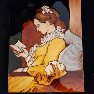 Puede incluir: Un azulejo tallado en madera que representa a una mujer con un vestido amarillo leyendo un libro mientras está sentada en una silla. El azulejo tiene un esquema de color marrón, amarillo y blanco.