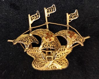 May include: A gold-tone metal brooch shaped like a sailing ship with intricate filigree wirework detailing. The ship features three masts with flags, curved sails, and small red cross emblems on the central and right sails, connected by thin wire.