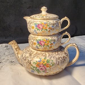 Puede incluir: Un juego de té vintage de tres piezas. El juego incluye una tetera, una azucarera y una crema, todas apiladas. Las piezas son de color crema con filigrana dorada y diseños florales en rosa, amarillo y azul. La tetera tiene un pico y un asa curvados.
