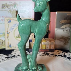 May include: A glossy, emerald green ceramic figurine of a standing fawn. The deer is depicted in profile, facing left, with its head turned slightly forward. It features a smooth, glazed finish and stands on a rectangular base.