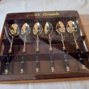 May include: A set of six gold-colored spoons with black handles, presented in a brown box. The box is labeled "Super Ceramic". The spoons have a classic design, suitable for serving. The spoons are in a clear plastic case.