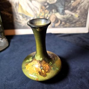 May include: A small, bulbous ceramic vase with a narrow neck and a flared rim. The vase features a dark olive green glaze with a hand-painted floral motif in shades of yellow and orange on the rounded body, showcasing a smooth, glossy finish.
