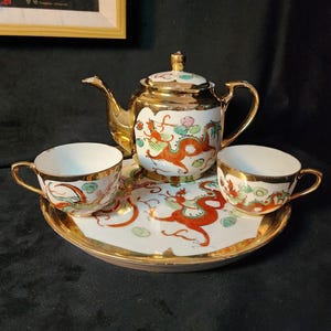 May include: A gold-edged porcelain tea set comprising a teapot, two teacups, and a serving tray. The set is adorned with a red dragon design and colourful details. The teapot has a lid and a curved spout.