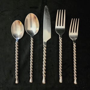 May include: A set of five silver-colored flatware pieces. The set includes two spoons, a knife, and two forks. Each piece has a twisted rope design on the handle. The flatware is arranged on a dark surface.