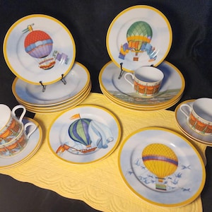 May include: A ceramic dinnerware set featuring hot air balloon illustrations. The collection includes plates and cups with yellow rims, white backgrounds, and colorful balloon motifs in red, green, blue, and yellow, set against a light blue sky.