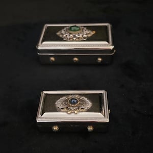 Puede incluir: Dos cajas rectangulares plateadas con adornos de joyas. La parte superior de cada caja es verde oscuro o negra con un diseño de joya central. La caja más grande tiene una joya verde, mientras que la más pequeña tiene una joya azul.