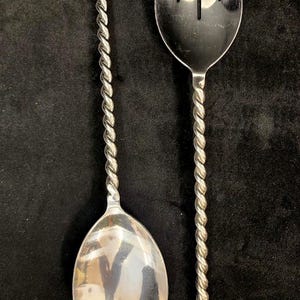 Puede incluir: Dos utensilios de servir plateados. Uno es una cuchara, el otro un tenedor. Ambos tienen un diseño retorcido en los mangos. La cuchara tiene un cuenco redondeado y el tenedor tiene tres púas. Los utensilios están sobre un fondo negro.