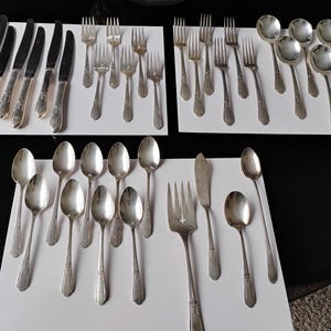 Op de afbeelding: Verzameling vintage zilveren bestek, waaronder messen, vorken, lepels en een botermes. Het zilverkleurige bestek is gerangschikt op witte oppervlakken tegen een donkere achtergrond, wat duidt op een formele dineropstelling.